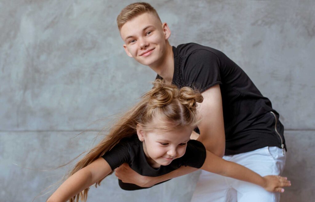 Deux enfants de la génération Alpha jouent ensemble, l’aîné simule un vol en tenant la plus jeune dans ses bras, exprimant complicité, énergie et imagination.